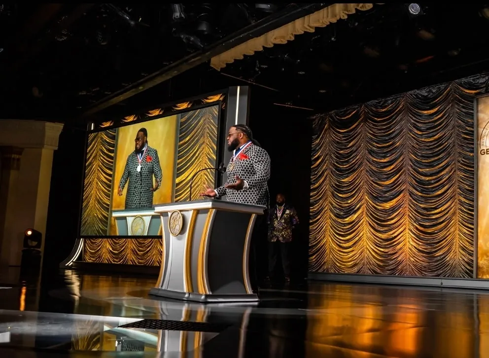 Taj Mahal speaking at a podium on stage, video reel